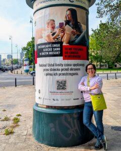 Edukatorka rodziców - Beata Broniek - stoi przy plakacie promującym chroniące zachowania rodziców w ramach akcji #warszawachroni